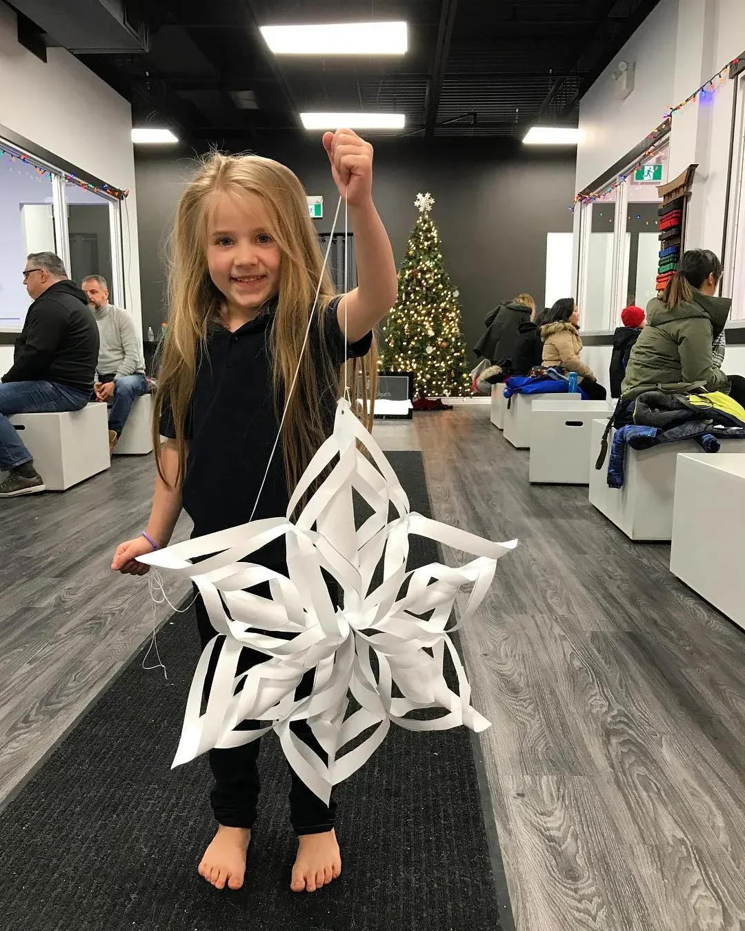 A girl holds up a big paper snowflake in the lobby, with a Christmas tree behind her