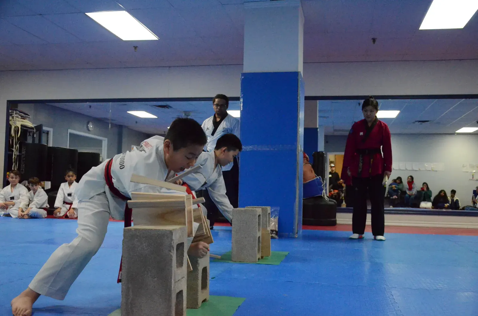 A student breaks boards set on blocks at a black belt test, as his instructors watch