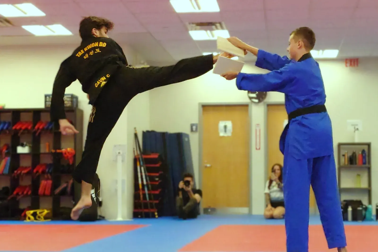 A black belt breaks a board with a jumping side kick while another black belt holds it