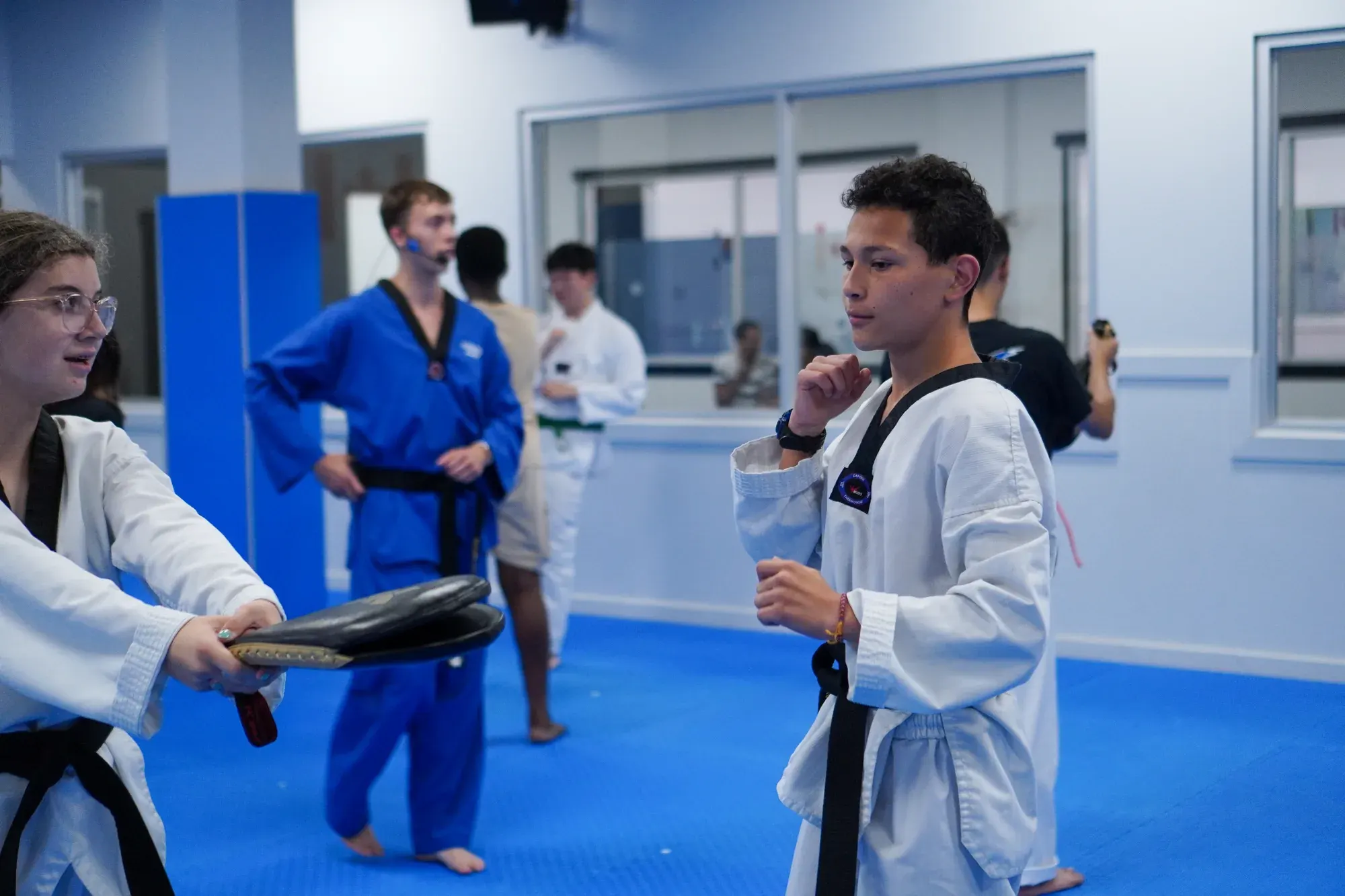 A black belt holds a paddle target for another black belt, with an instructor in a blue uniform behind them