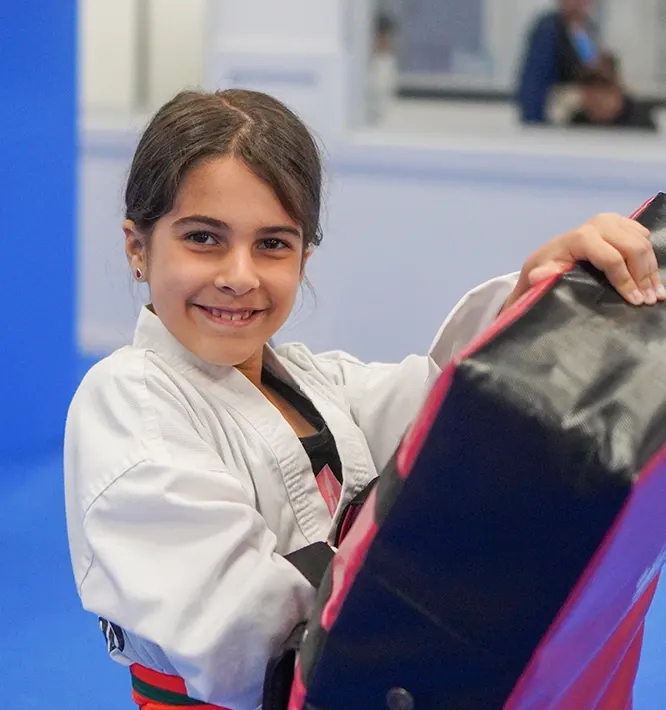 A smiling student in a white uniform holds a kick pad