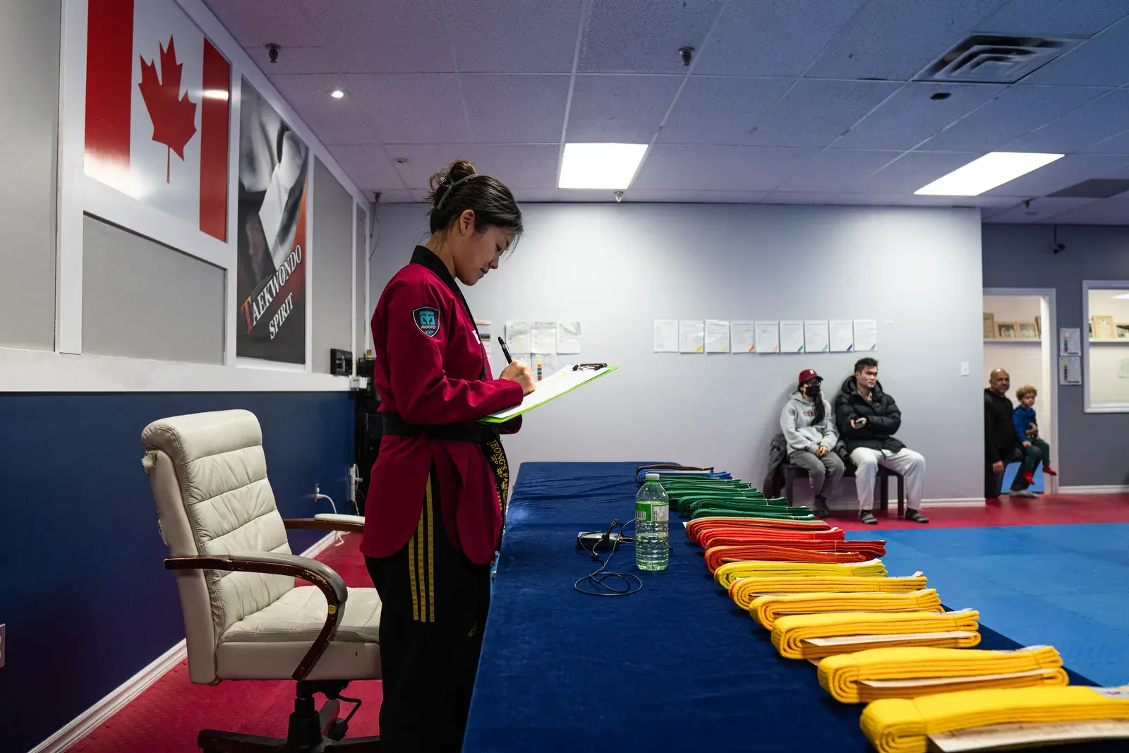 An instructor holds a clipboard at a belt test, with new belts laid out on the table
