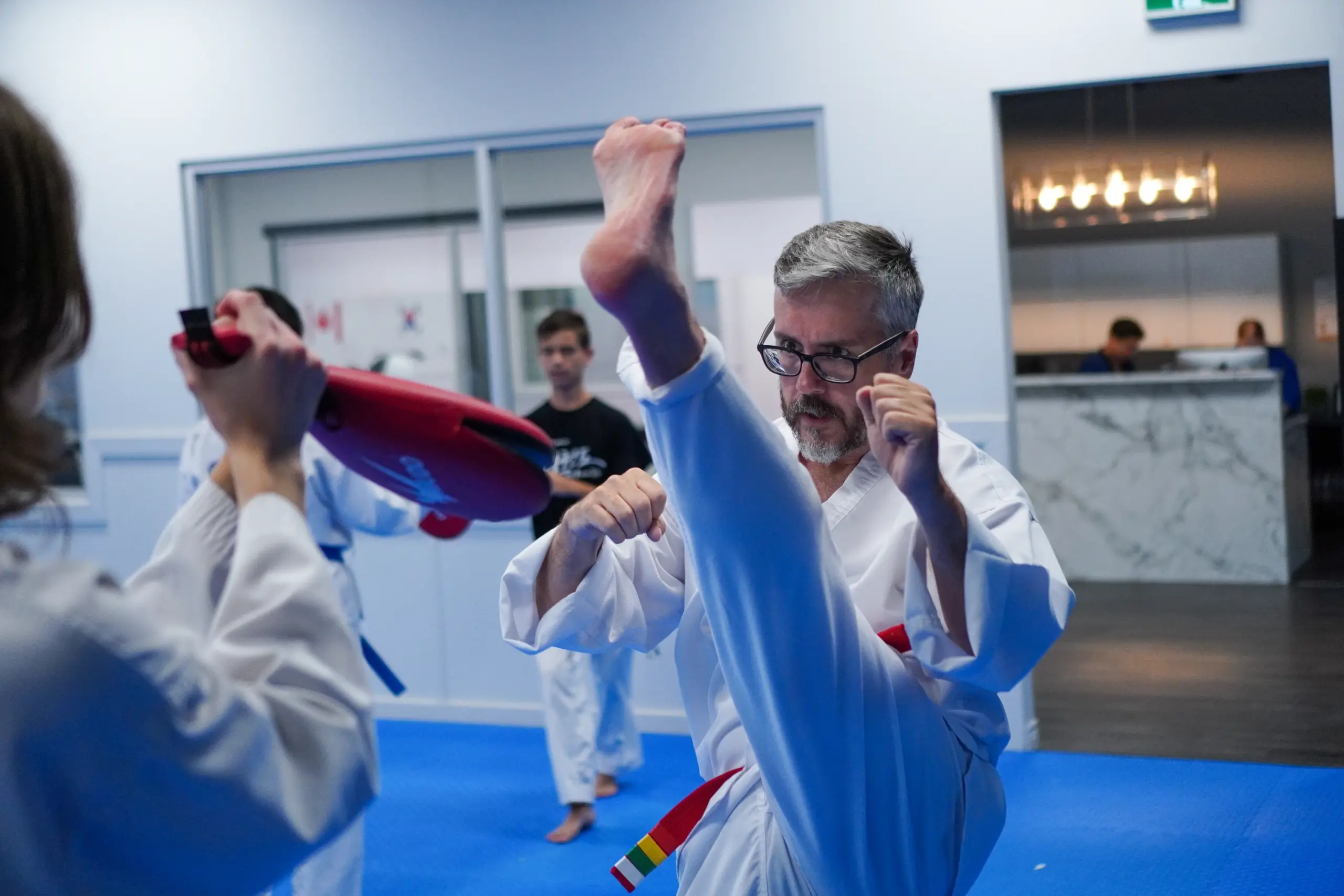 An adult student with grey hair and glasses throws a high kick beside a partner holding a pad