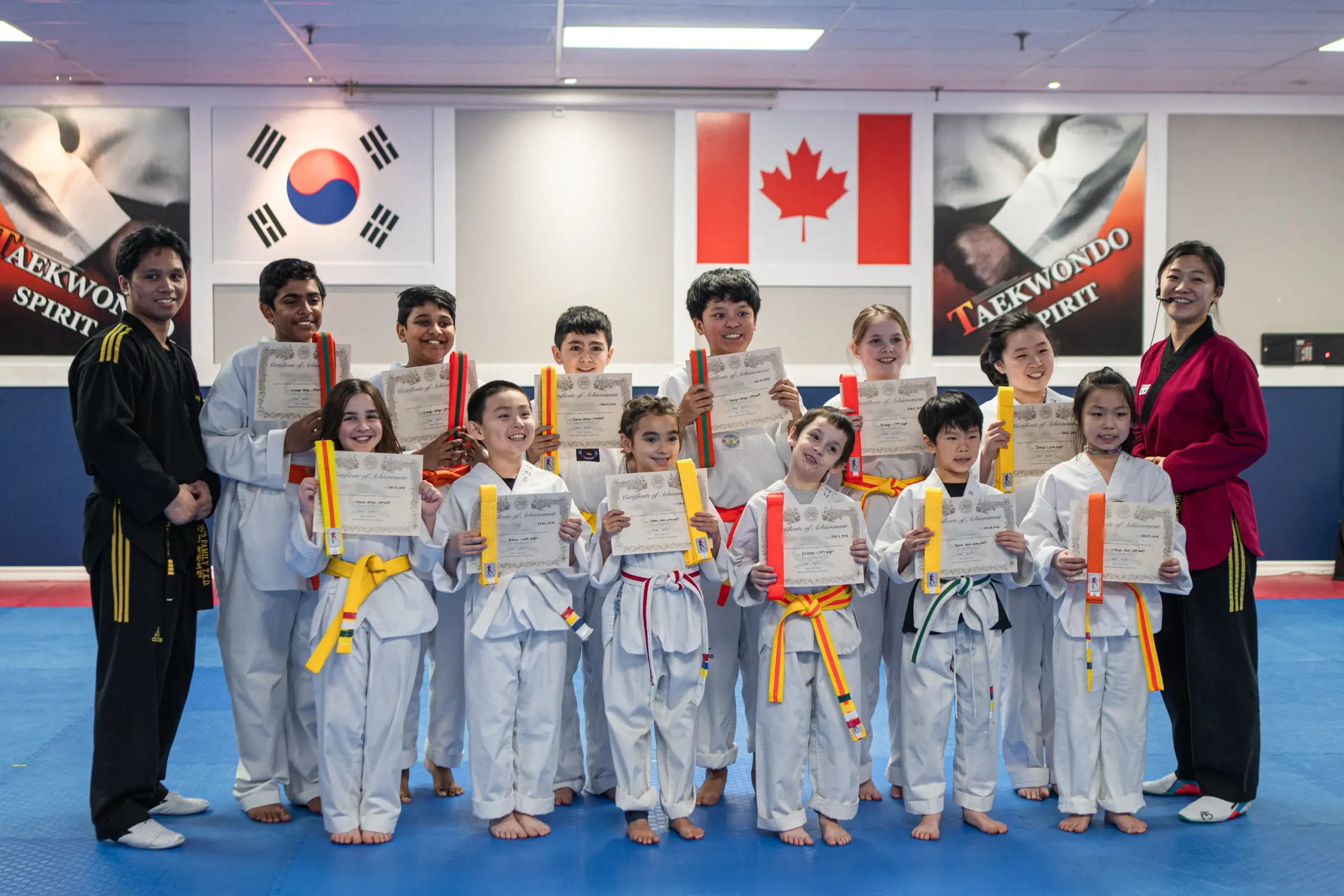 Smiling students hold up their new belts and certificates with two instructors, under the Korean and Canadian flags