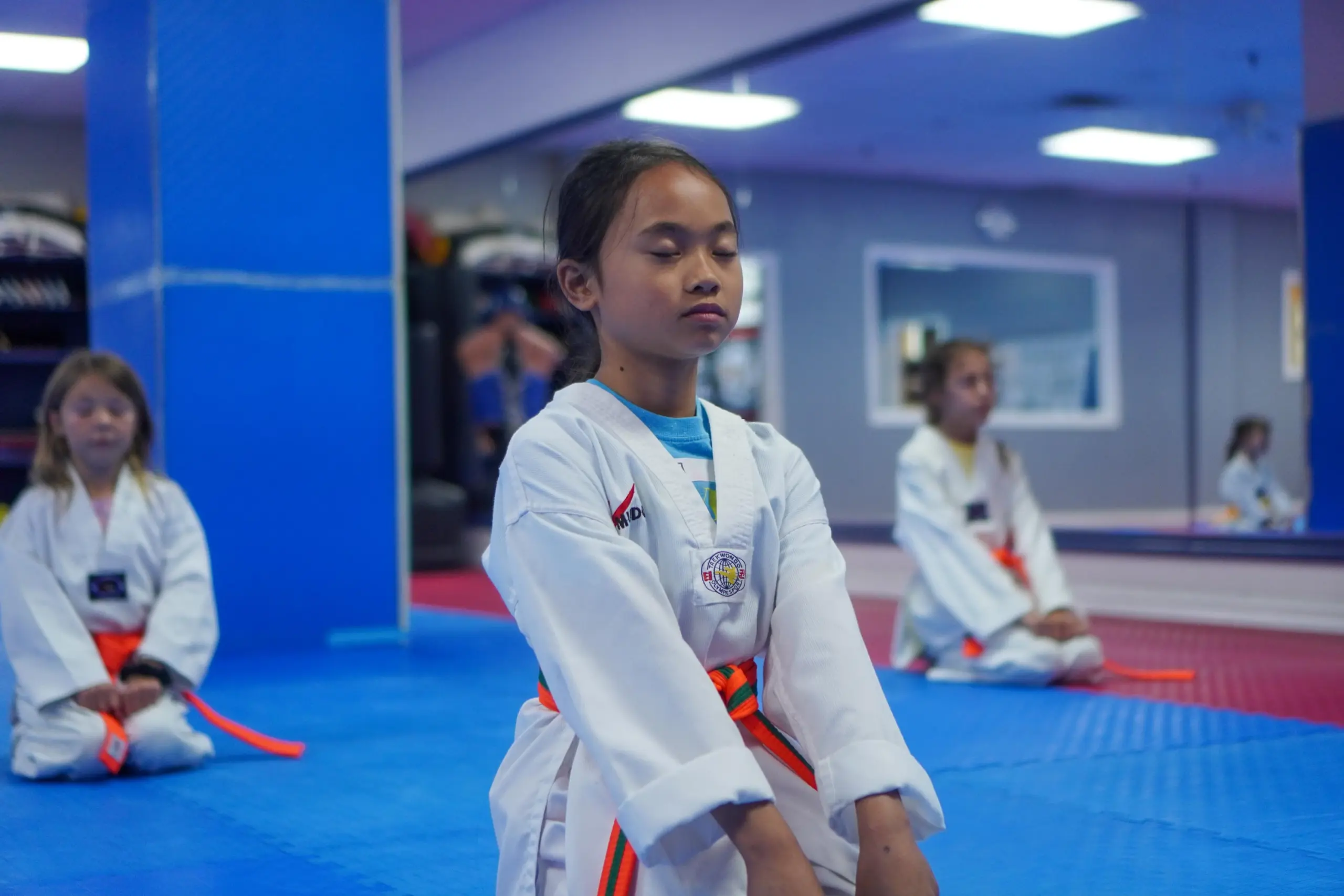 A young student in an orange belt kneels with her eyes closed, with her classmates behind her