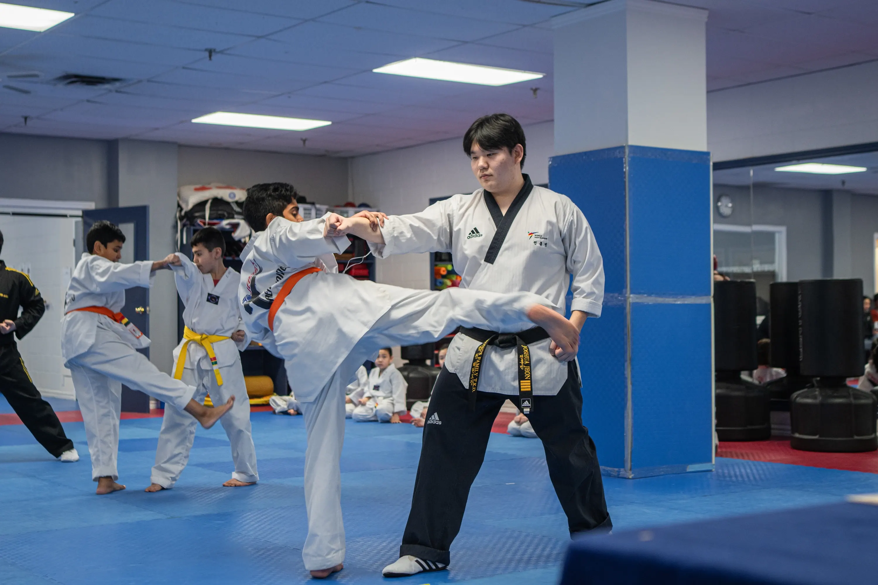 A student in an orange belt kicks toward his instructor, with classmates practising kicks behind them