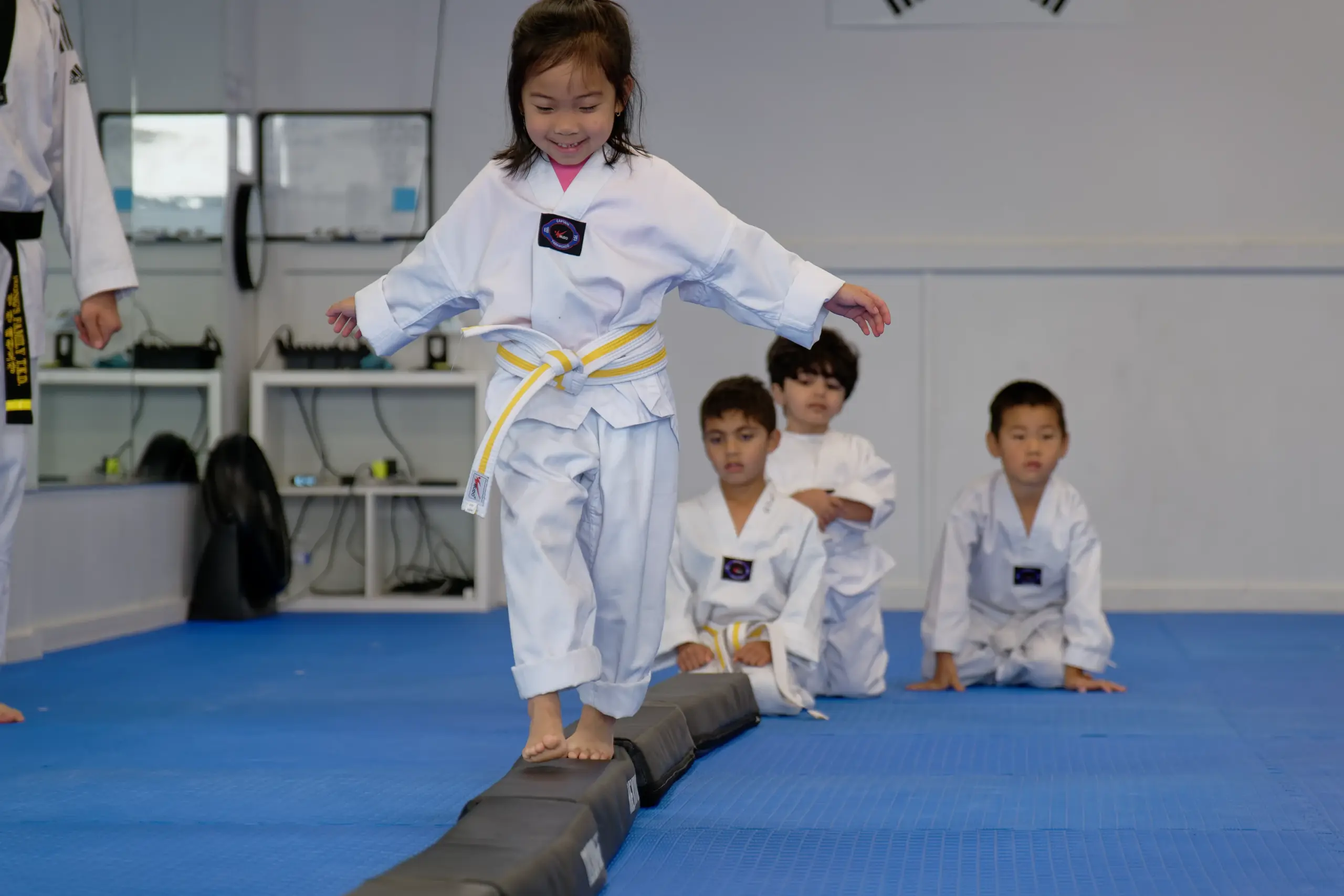 A smiling young student in a yellow belt walks along a balance beam while her classmates wait their turn