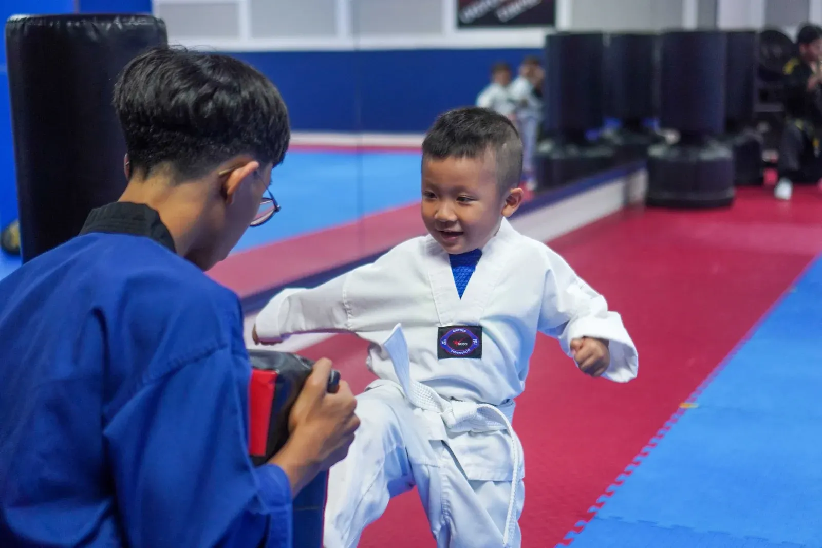 A smiling young student in a white belt kicks a pad held by his instructor